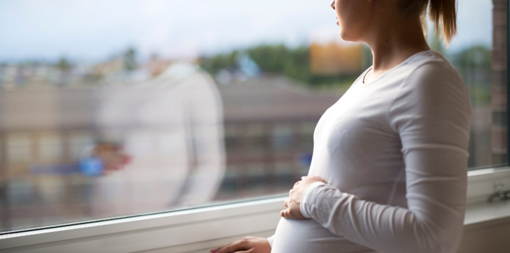 Pregnant woman looking out a window while holding her belly.