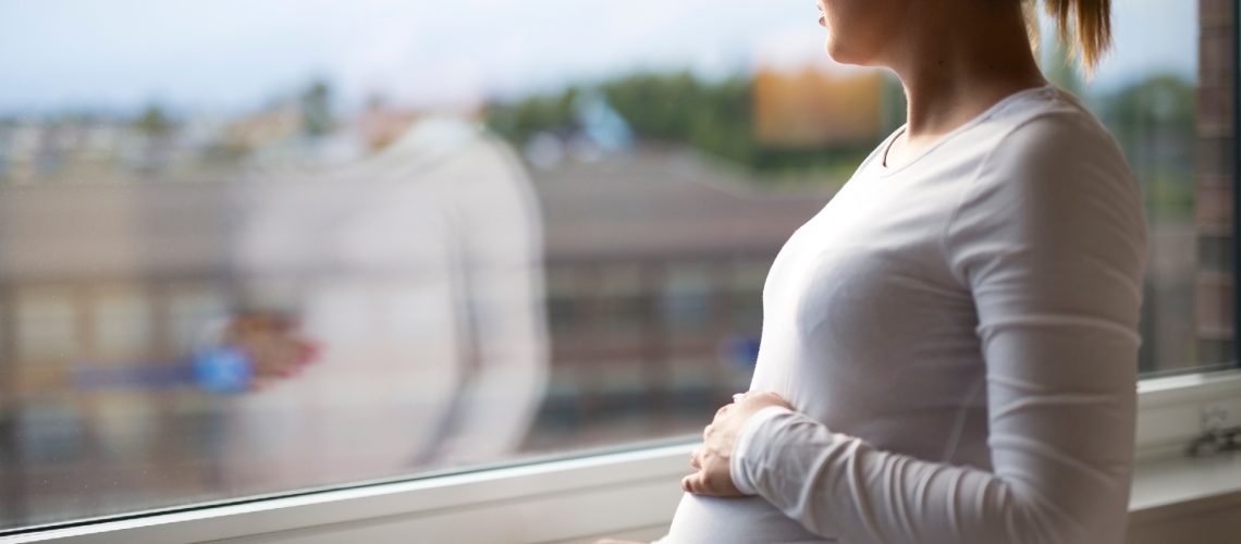 Pregnant woman looking out a window while holding her belly.